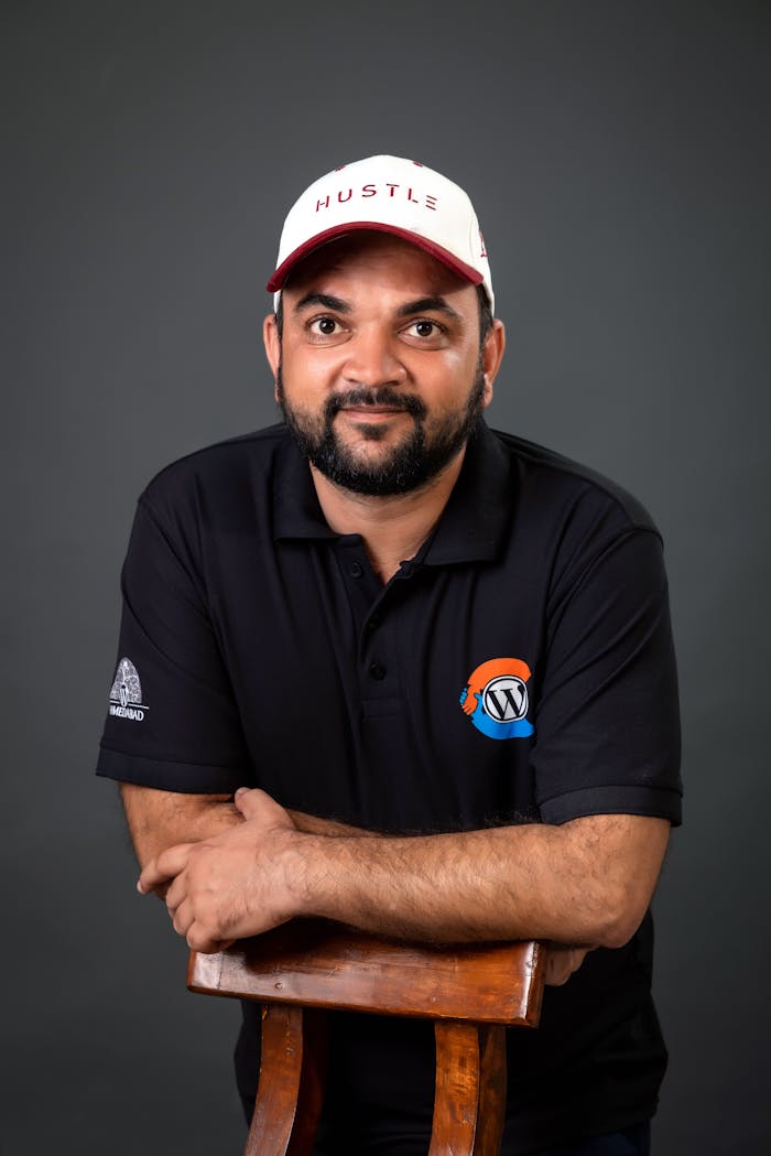 Smiling man in a black polo with logos and cap standing indoors against a dark background.